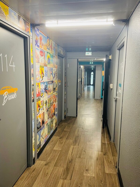 Un couloir doté d’un plancher en bois, de portes numérotées et d’un mur décoré d’affiches colorées.