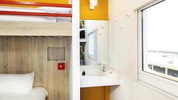 Un lit superposé avec un panneau décoratif en bois, un lavabo blanc avec un miroir au-dessus, et une fenêtre donnant sur un parking.