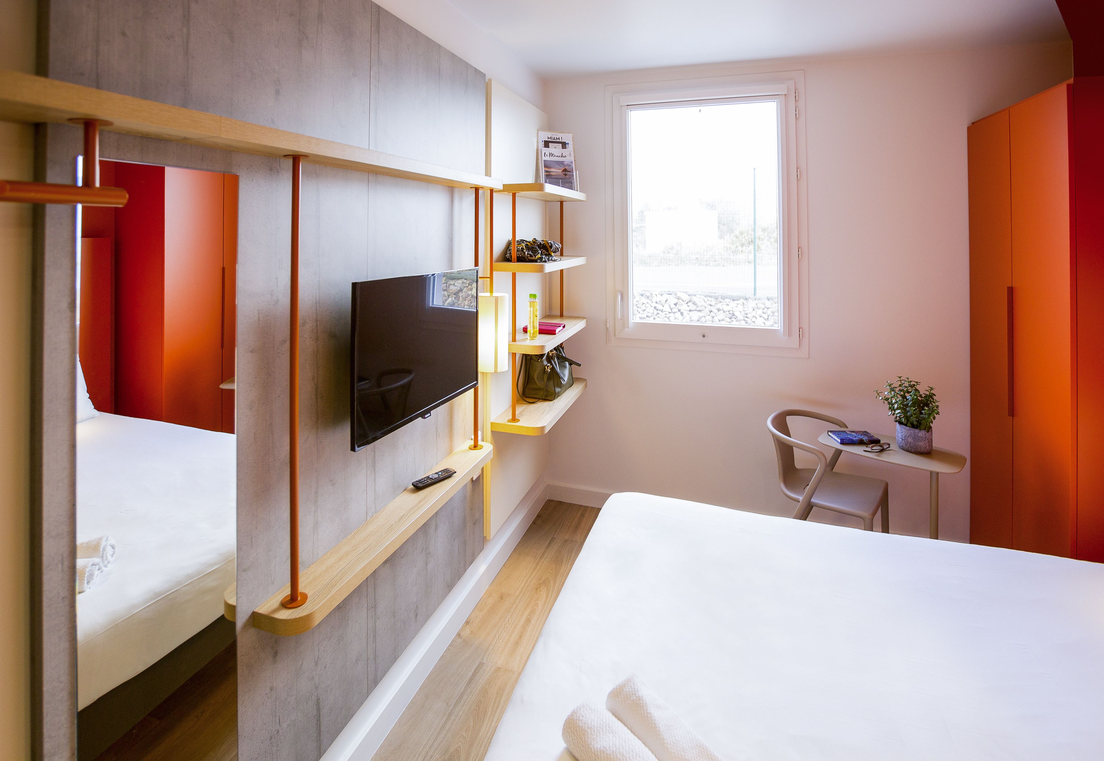 Habitación de hotel moderna con cama, televisión montada en la pared, una mesita con una planta y una ventana que proporciona luz natural.