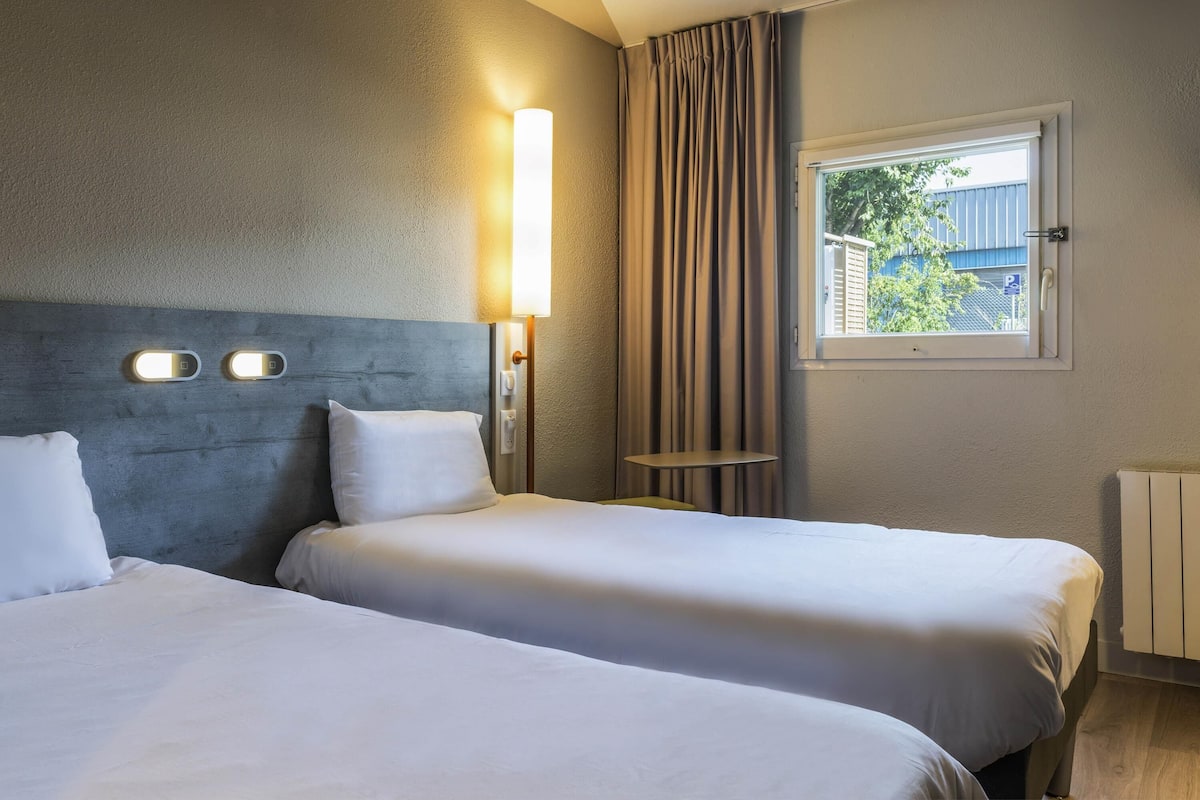 Une chambre d’hôtel avec deux lits, une table de chevet, une lampe, une fenêtre avec des rideaux et une vue sur la verdure.