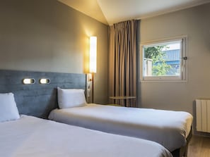 Ett hotellrum med två sängar, ett nattduksbord, en lampa, ett fönster med gardiner och utsikt över grönområden utanför.