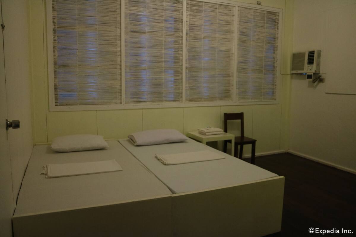 A room with two beds, a small table, a chair, and a window with blinds.