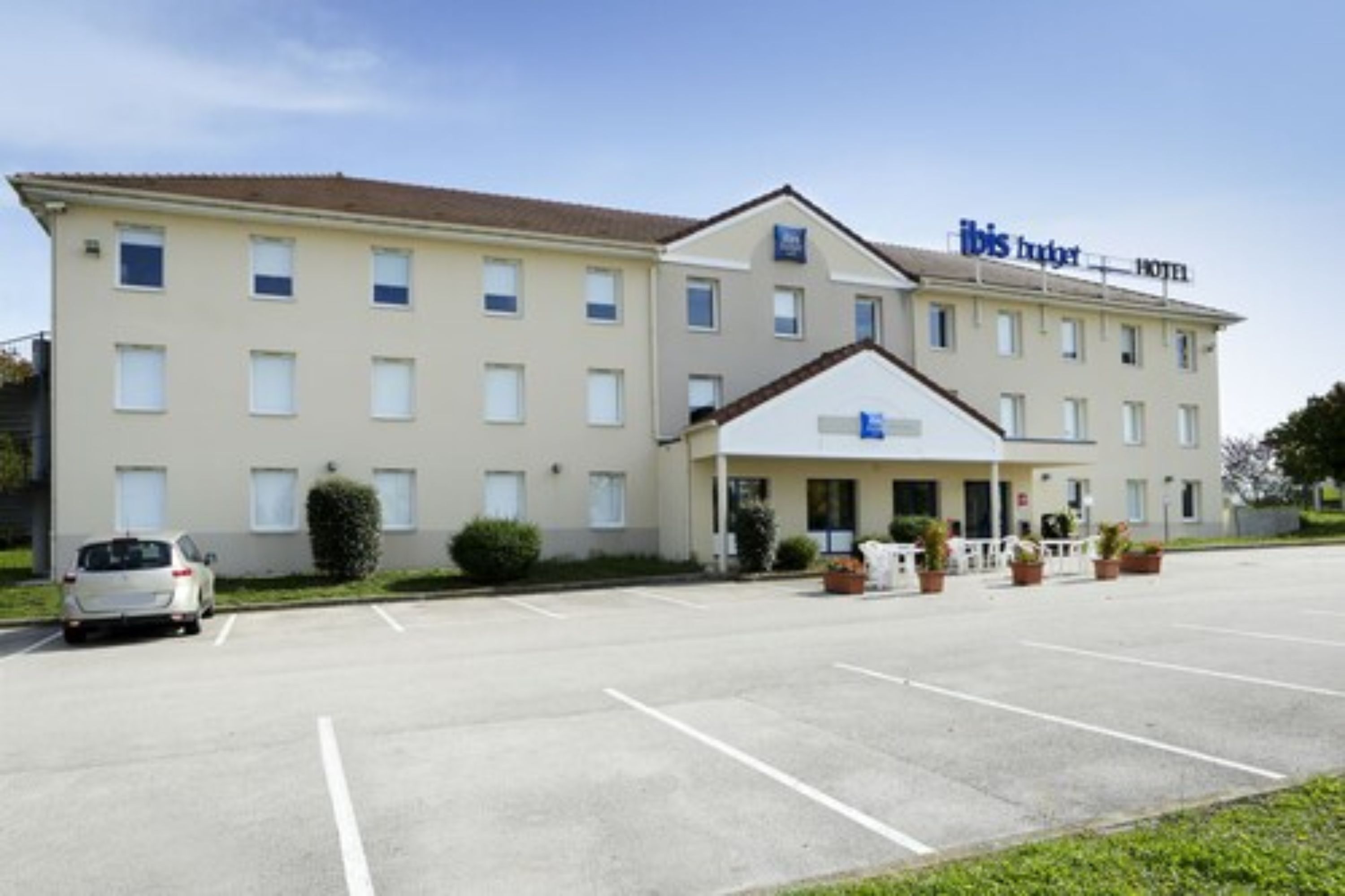 Ein zweistöckiges Hotel mit flachem Dach, einem Schild mit der Aufschrift 'ibis budget HOTEL' und einem Parkplatz mit einigen Autos.