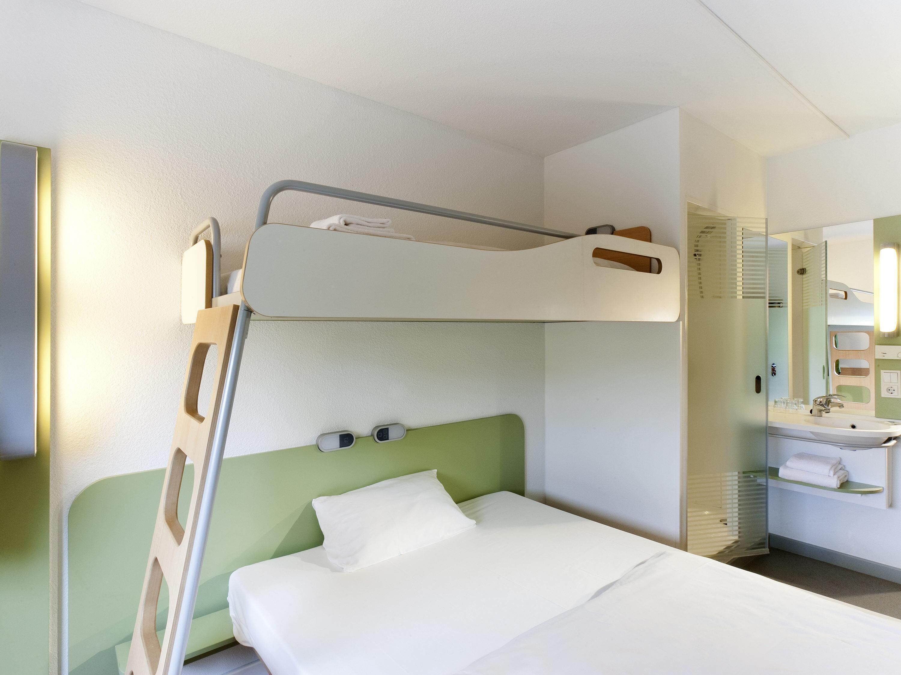 A bunk bed with a headboard, a bedside table, a mirror, and a bathroom with a sink and towel rack.