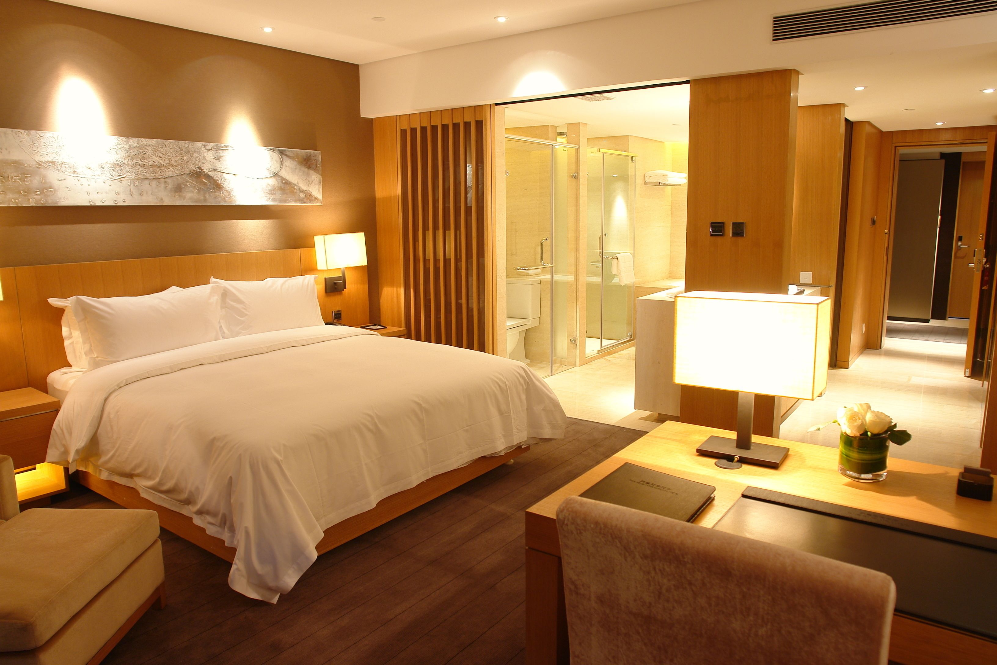 A hotel room with a large bed, bedside tables, a desk with a lamp, a chair, and a shower area visible through a glass door.