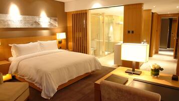 A hotel room with a large bed, bedside tables, a desk with a lamp, a chair, and a shower area visible through a glass door.