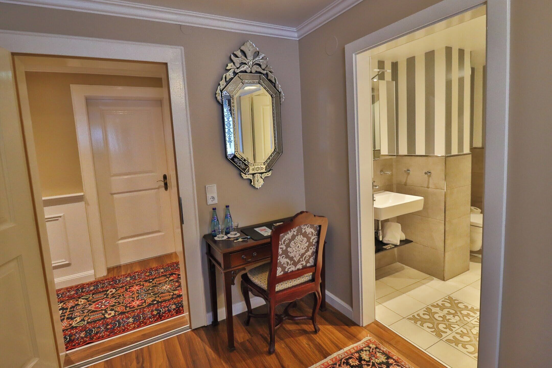 A room with a wooden desk, a chair, a mirror, and a bathroom with a sink and toilet.