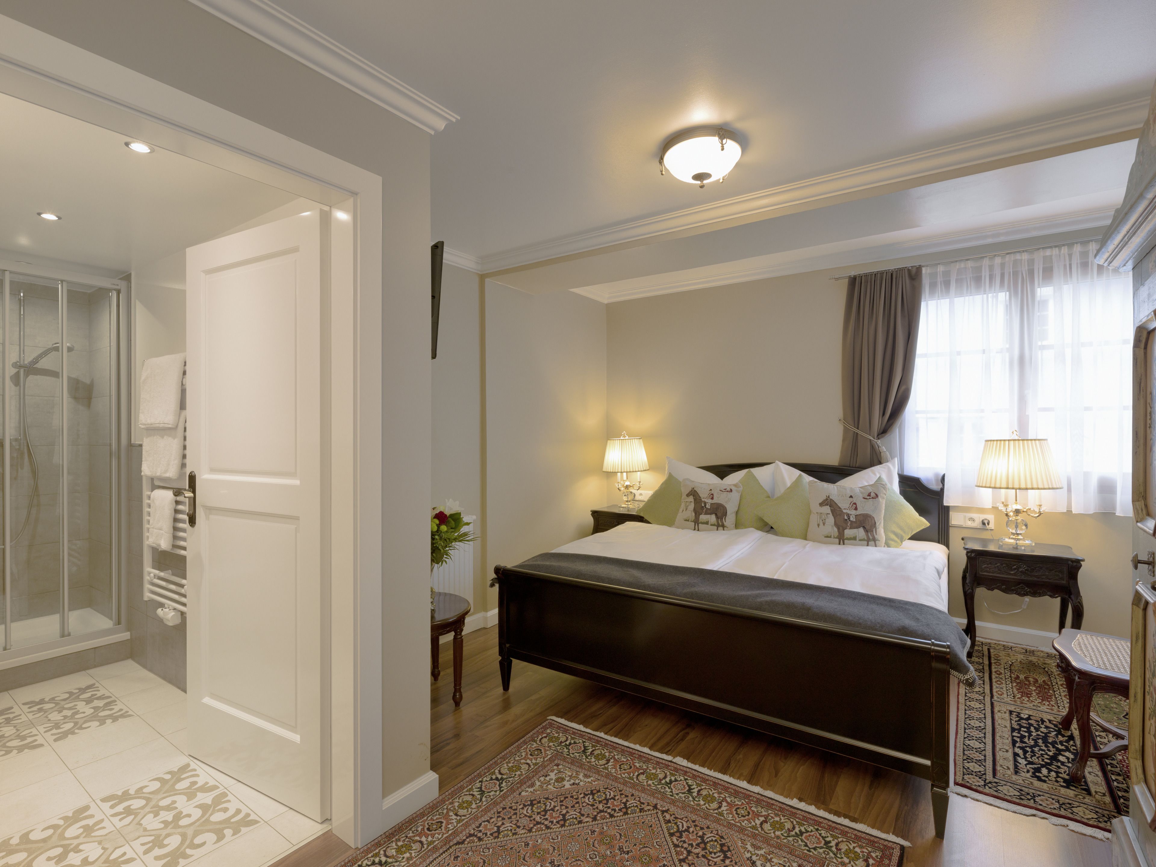 Une chambre avec un grand lit, une table de chevet avec une lampe, une commode, une fenêtre avec des rideaux et une salle de bain avec une douche et un porte-serviettes.