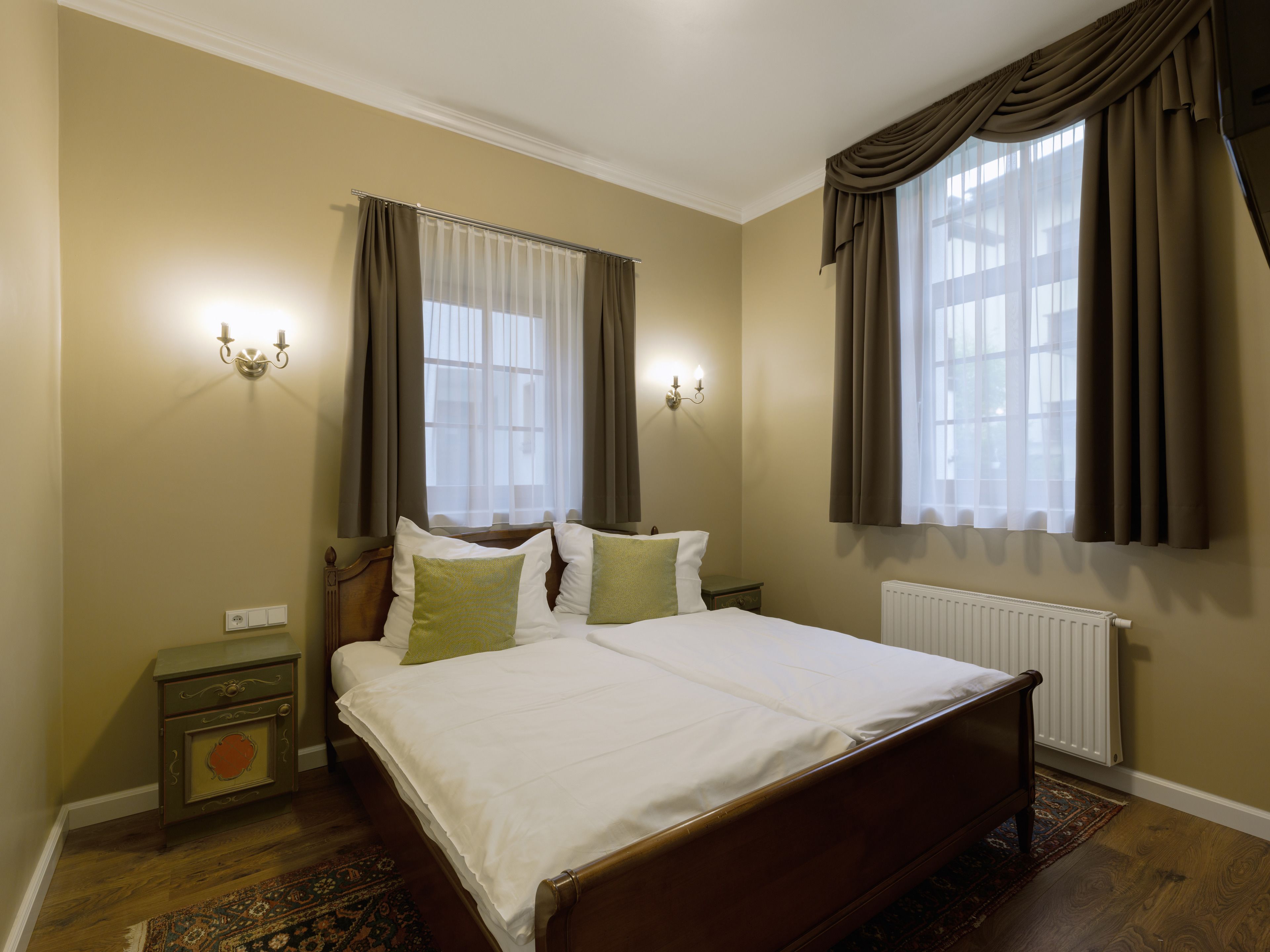A bedroom with a large bed, two bedside tables, a nightstand, a radiator, and two windows with curtains.