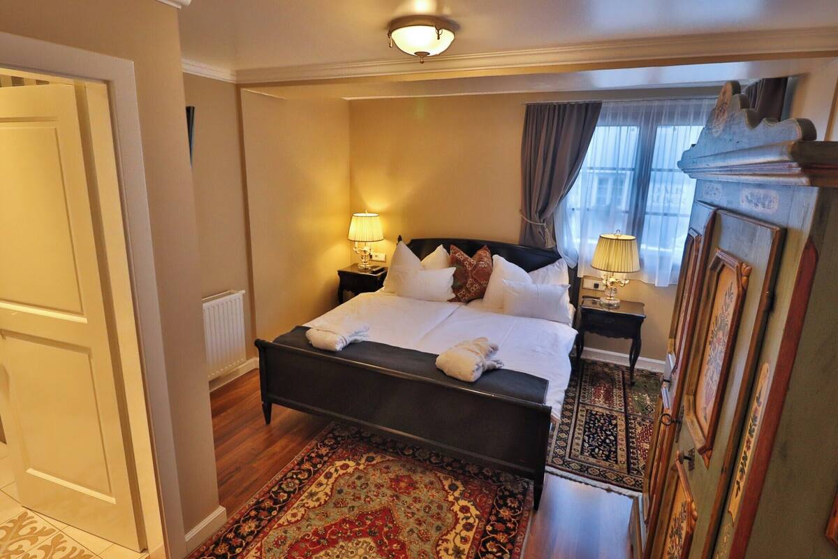 A hotel room with a bed, two bedside lamps, a patterned rug, a radiator, and a wooden wardrobe.
