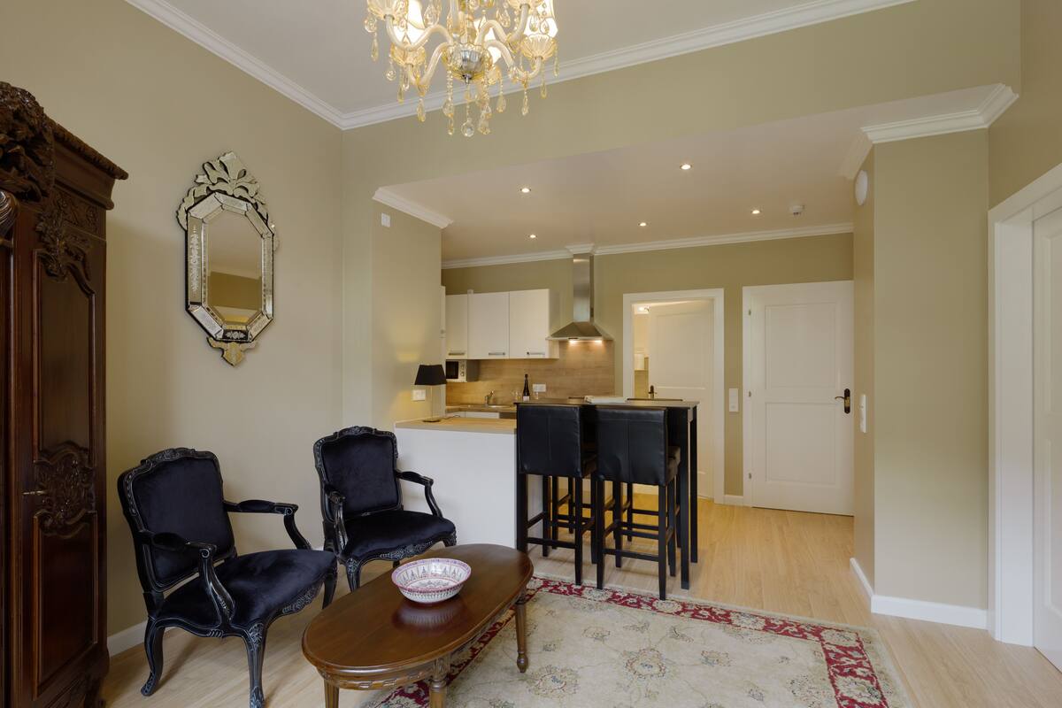 A dining area with a wooden table, two chairs, a chandelier, and a kitchen in the background.