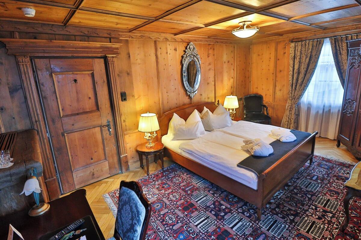 A wooden-paneled room with a large bed, two bedside lamps, a mirror, and a patterned rug.