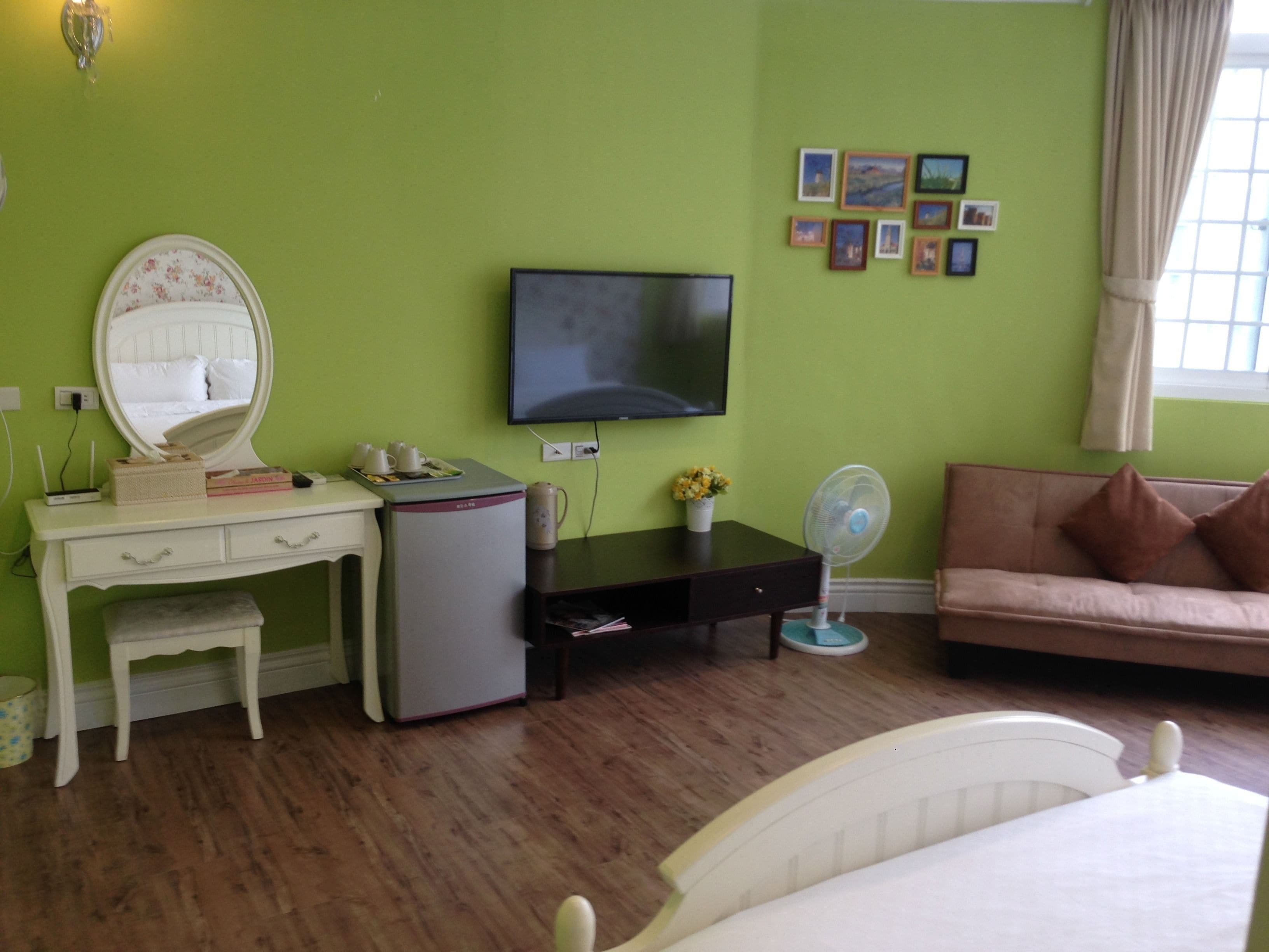 Una habitación con pared verde, cama, escritorio con espejo, televisor, ventilador y sofá.