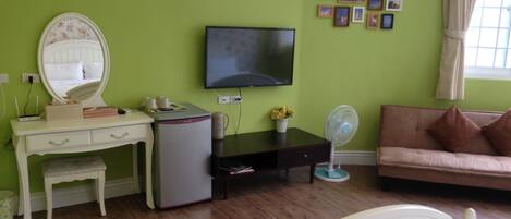 Una habitación con pared verde, cama, escritorio con espejo, televisor, ventilador y sofá.