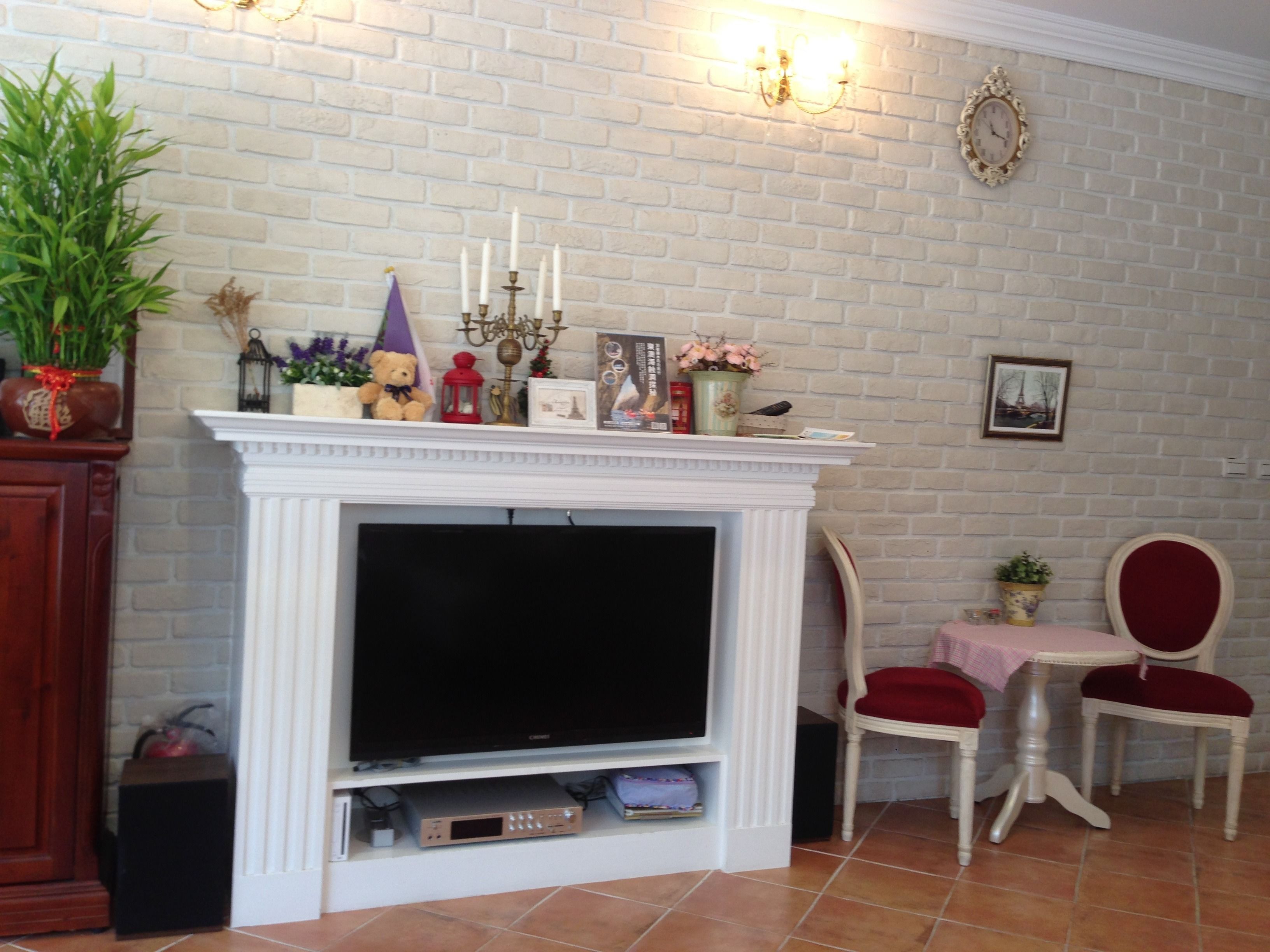 Una sala de estar con chimenea de ladrillo blanco, un televisor y dos sillones tapizados en rojo.