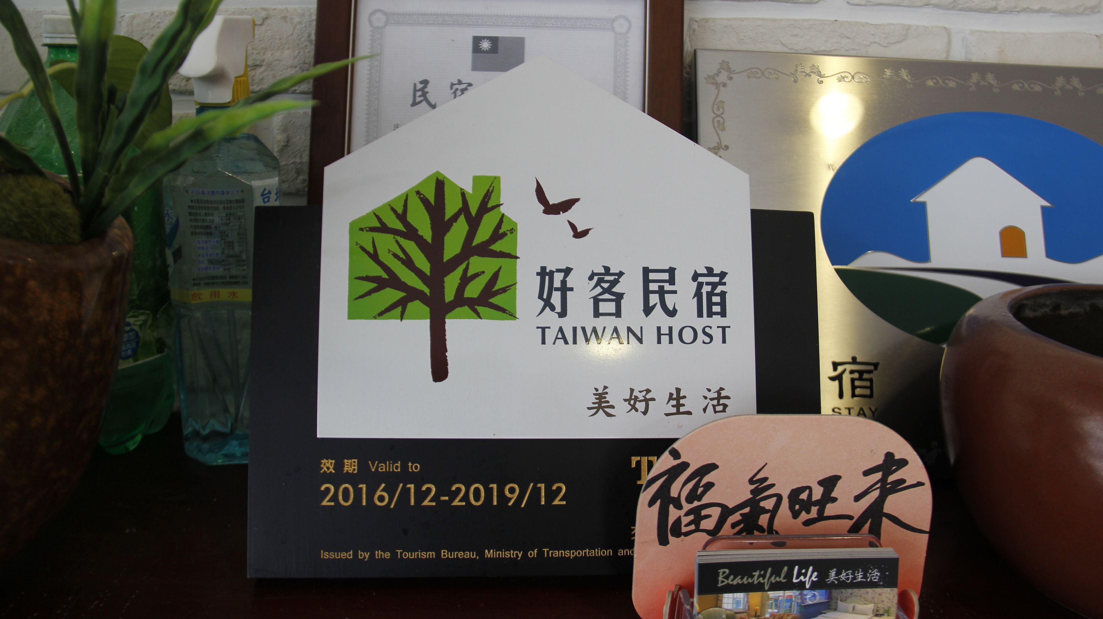 Un senyal amb una il·lustració d'un arbre i un ocell, el text 'Taiwan Host' i 'Vida Bonica', i un rang de dates de '2016/12-2019/12'.