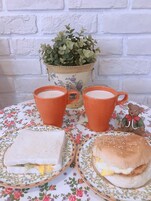 Una mesa con mantel floreado, dos tazas anaranjadas, un sándwich, una hamburguesa y una pequeña figurita de un oso.