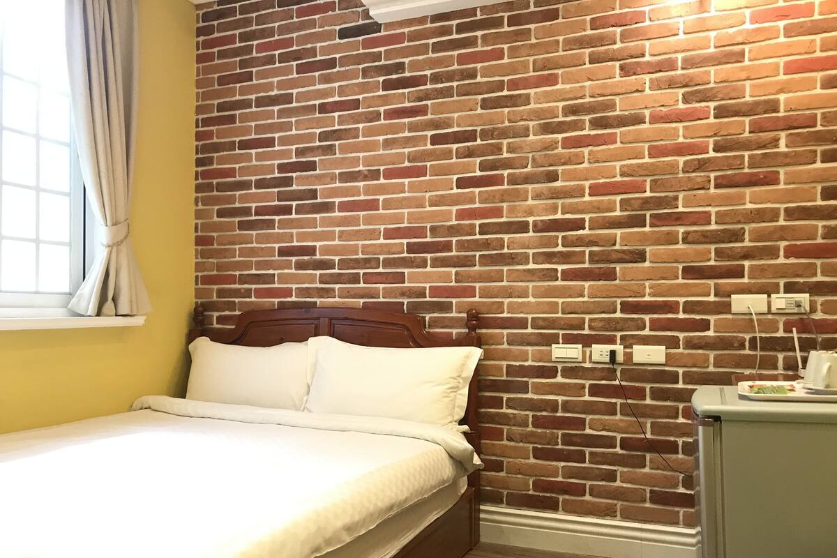 A bedroom with a brick-patterned wall, a wooden bed frame, a bedside table with a lamp, and a window with curtains.