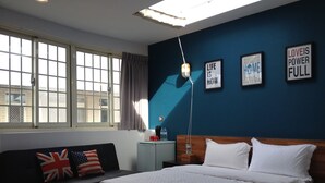 Habitación de hotel moderna con una cama grande, un sofá con banderas británica y estadounidense, una mesita de noche, una lámpara y un cuadro motivacional en la pared.