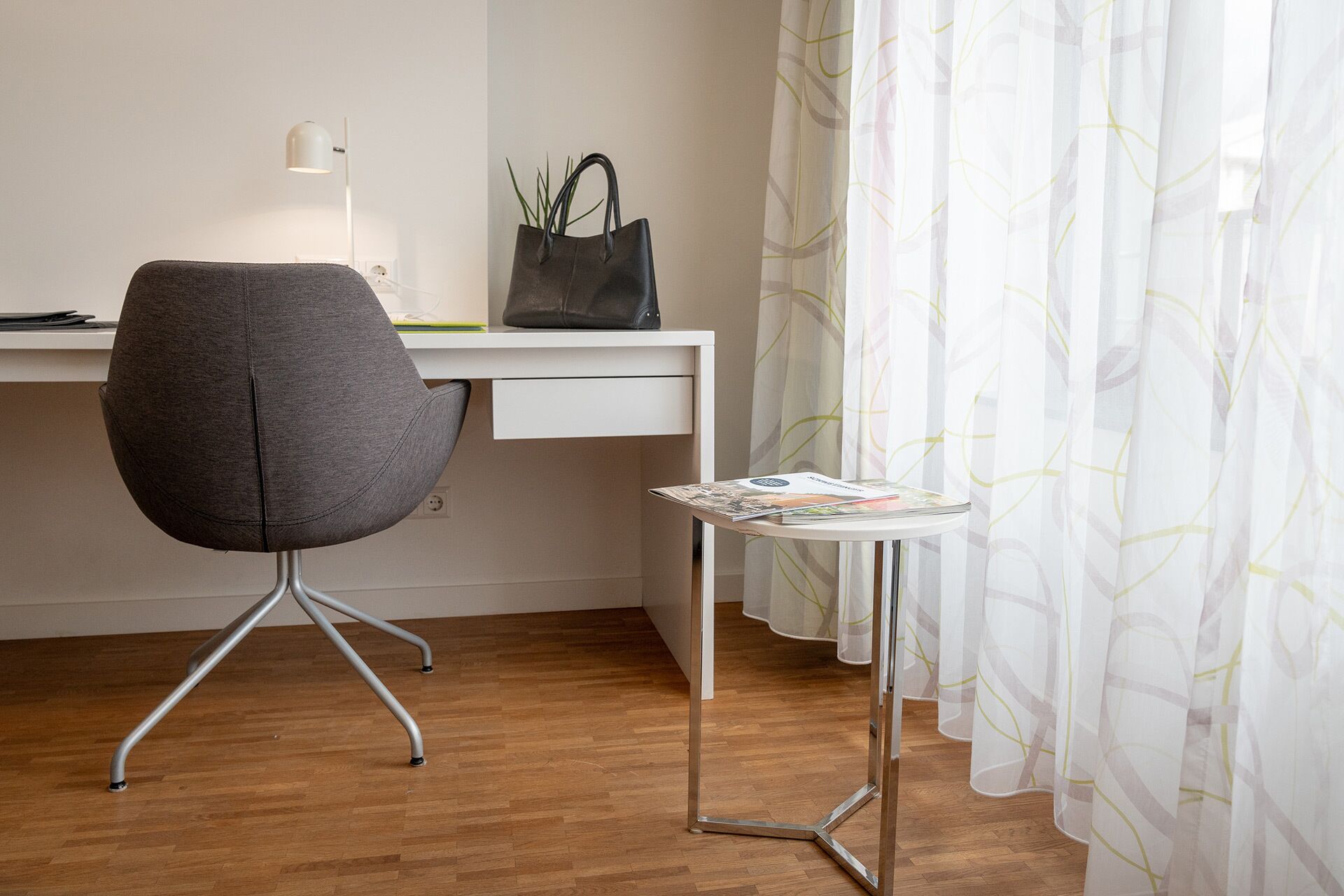 A modern office space with a grey office chair, a white desk, and a small side table with a magazine.