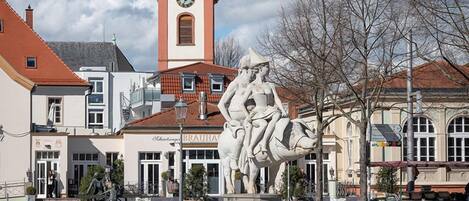 A public square with a central sculpture, a clock tower, and surrounding buildings.