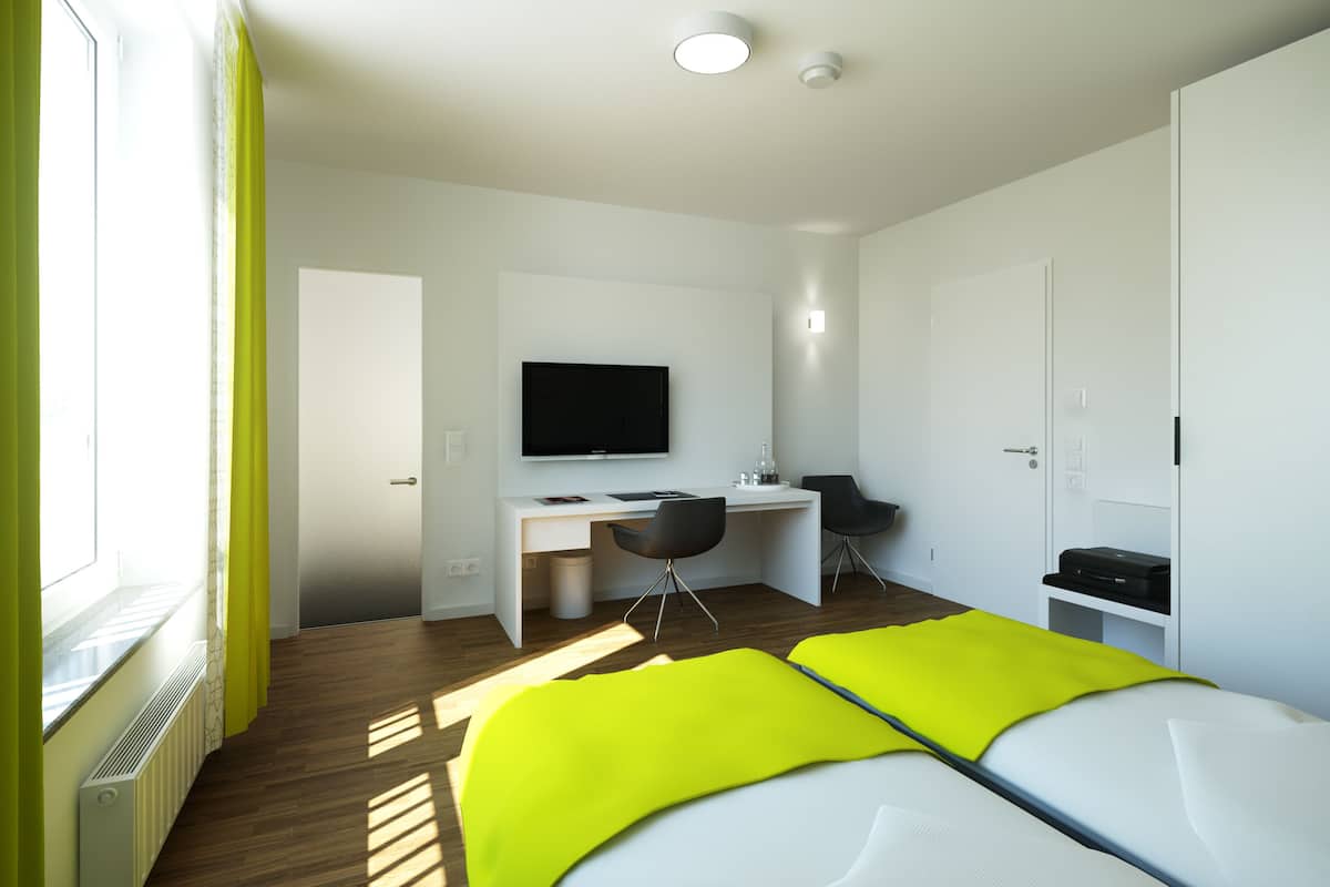 A modern hotel room with a bed, a desk, a chair, a television, and a wardrobe.