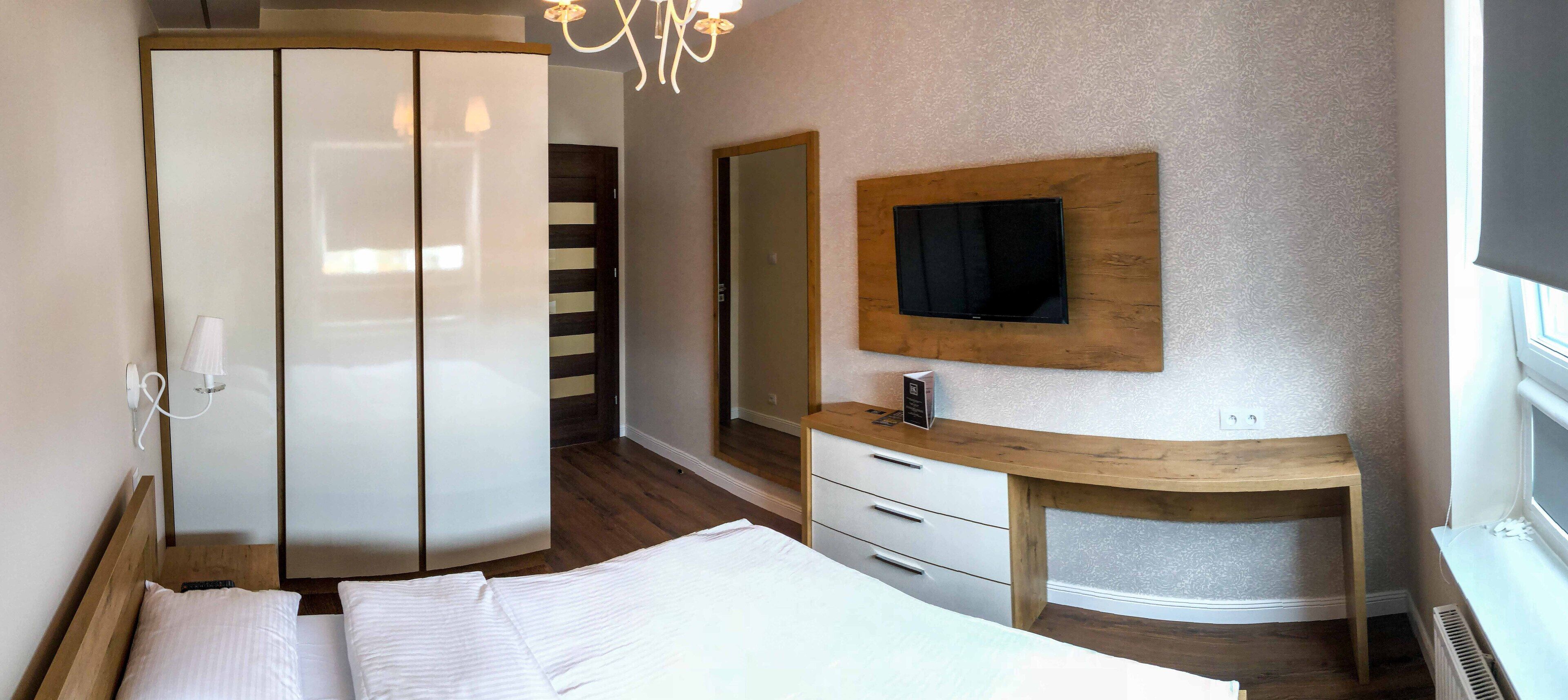 A bedroom with a bed, a wooden desk, a mirror, and a television mounted on the wall.