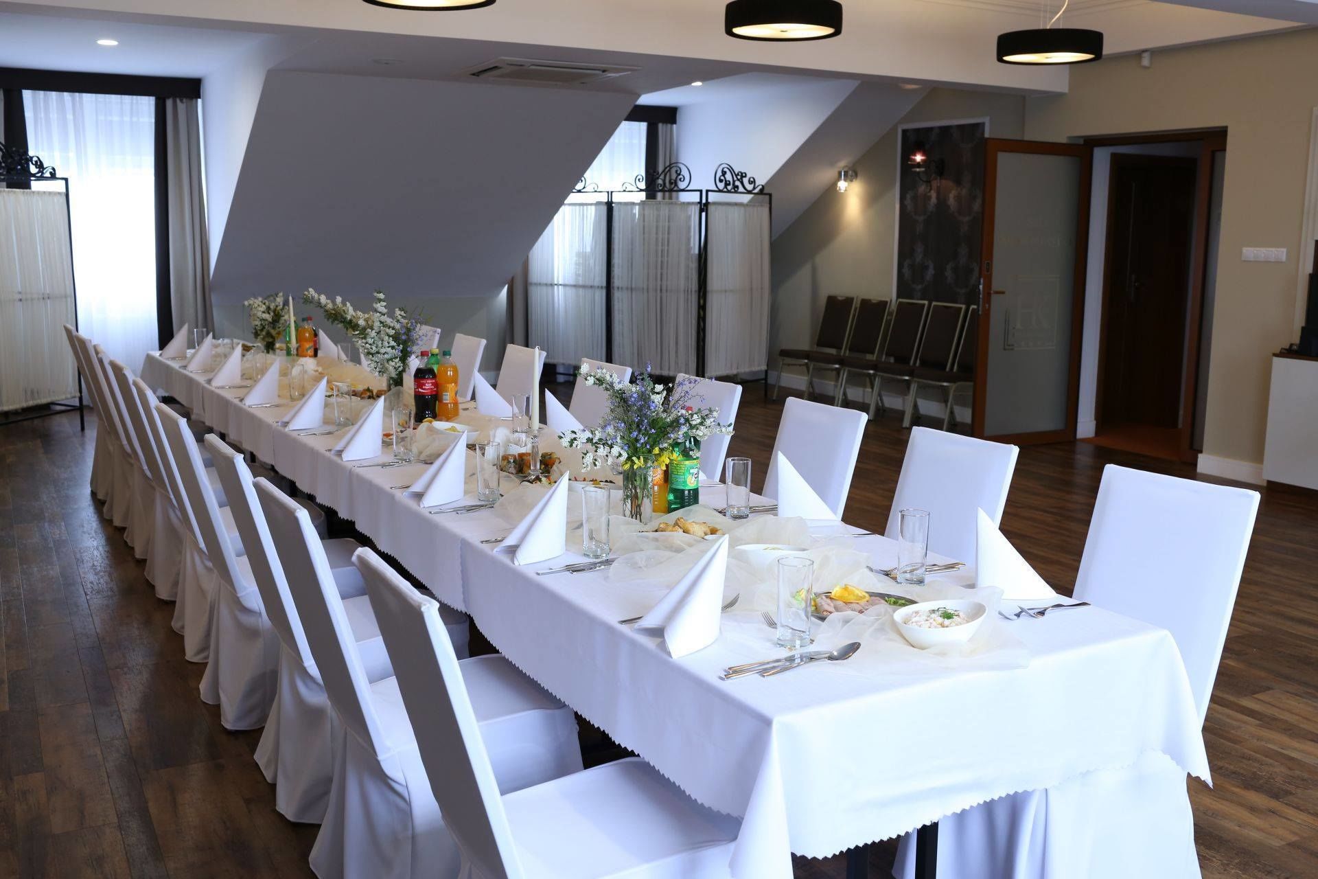 A long dining table set for a formal event, with white tablecloths and chairs, in a room with a modern design and a view of a staircase.