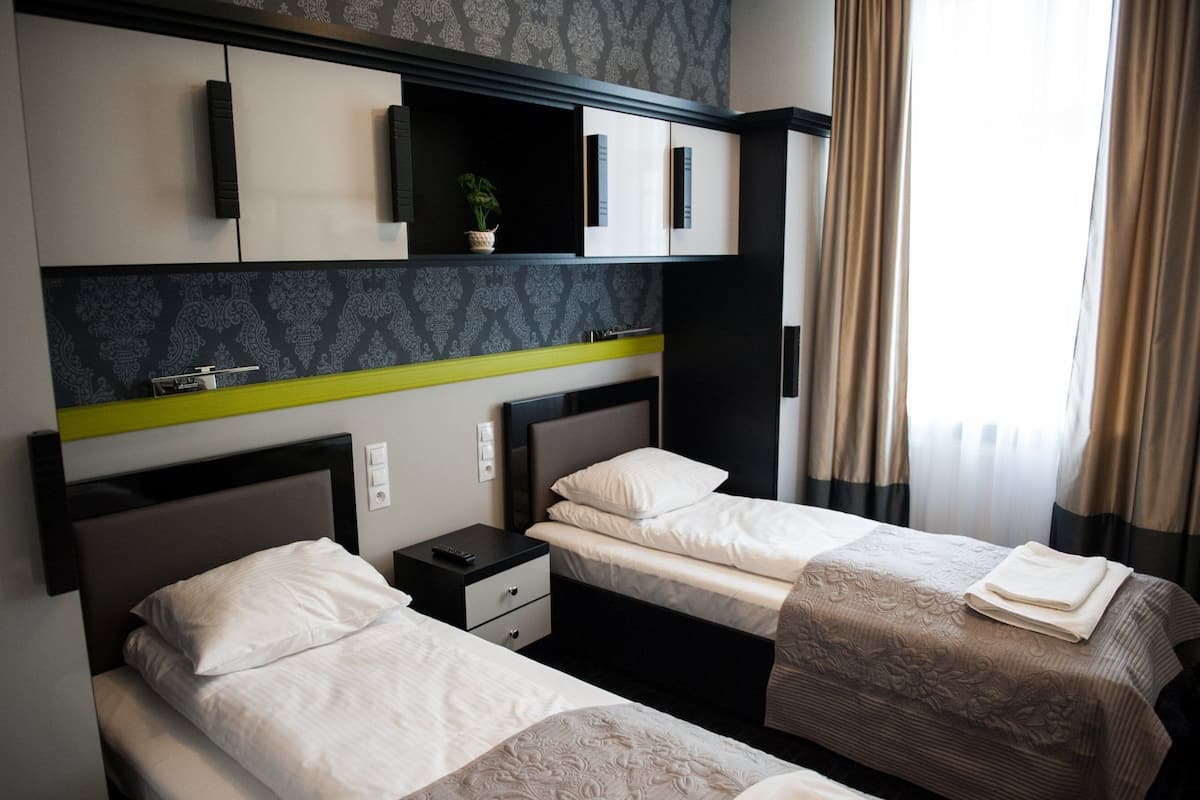 A hotel room with two beds, a nightstand, a shelf with a plant, and a window with curtains.