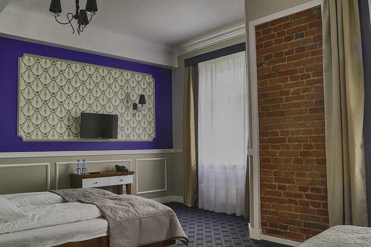 A hotel room with a bed, a bedside table with a lamp, a television, and a brick wall.