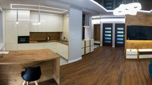 Una cocina moderna con armarios blancos, encimera de madera y microondas integrado. Hay una zona de comedor con mesa de madera y una silla negra. El cuarto tiene piso de madera y un sofá azul.