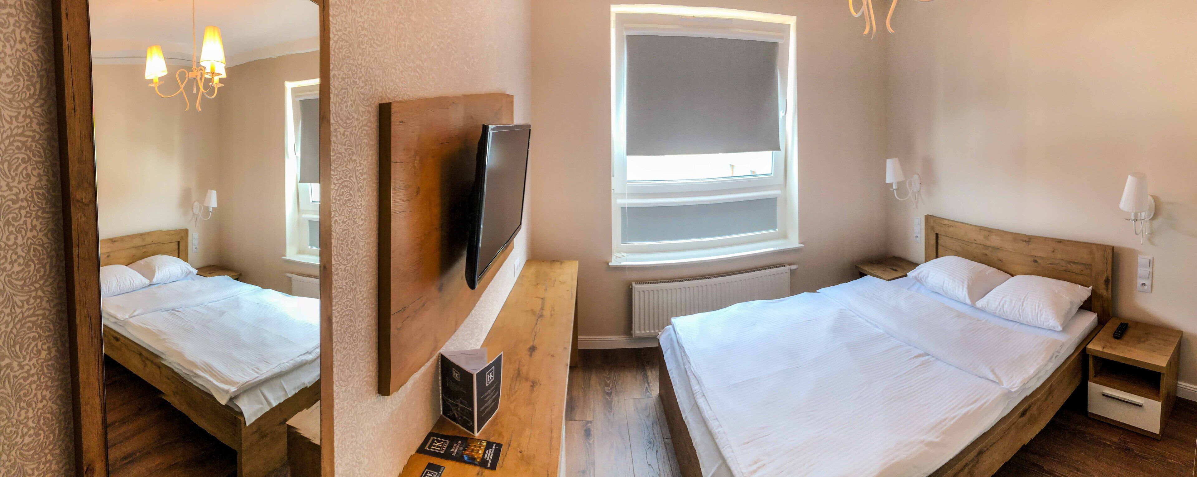 A double bed with a wooden headboard, a bedside table, a nightstand, a TV mounted on the wall, and a window with a blind.