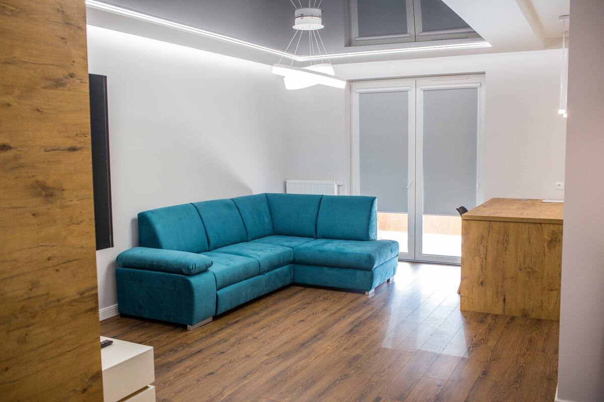 A modern living room with a teal sectional sofa, wooden flooring, a wooden dining table, and a sliding glass door leading to a balcony.