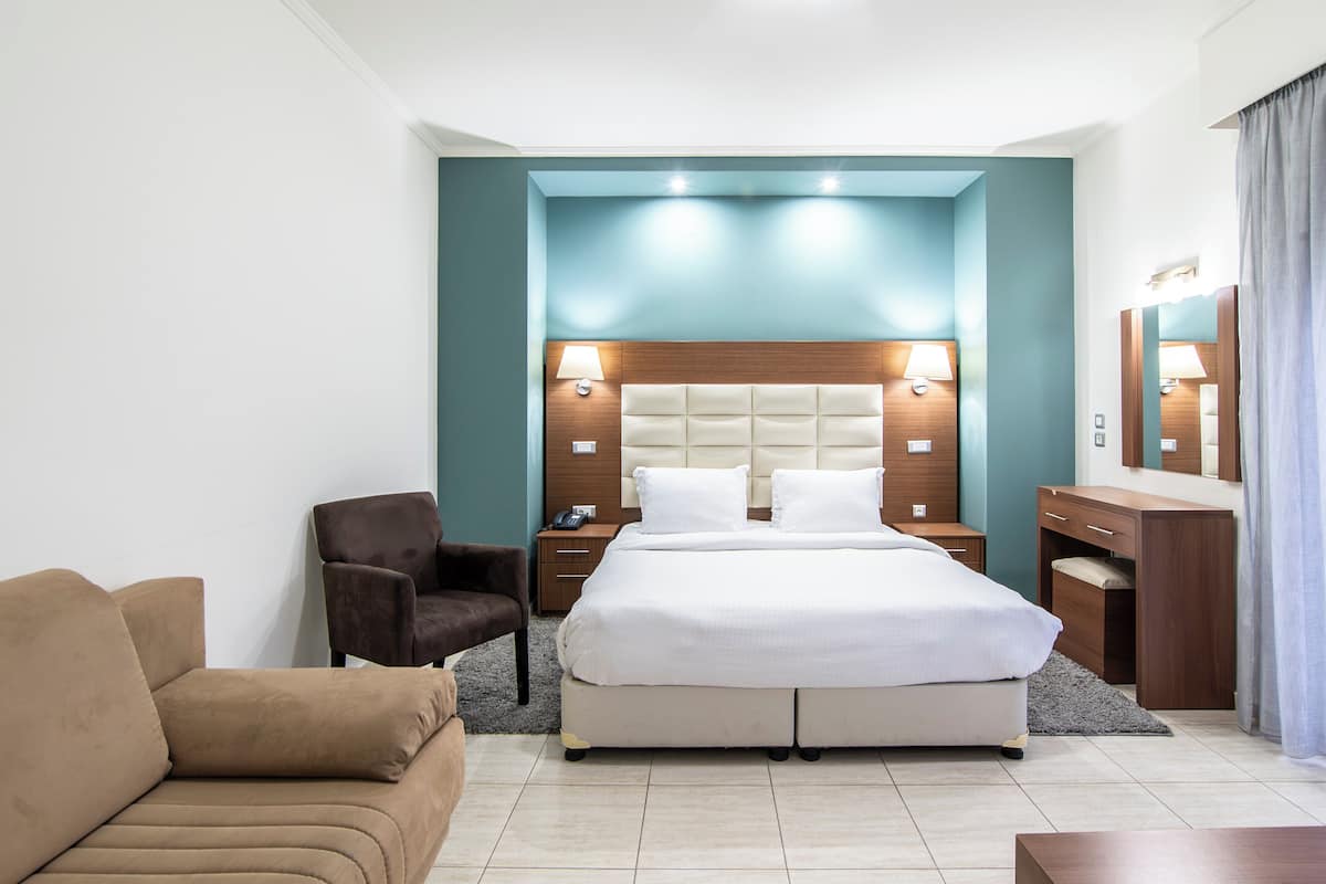 Une chambre d’hôtel avec un grand lit, un fauteuil marron, un bureau en bois et un miroir.