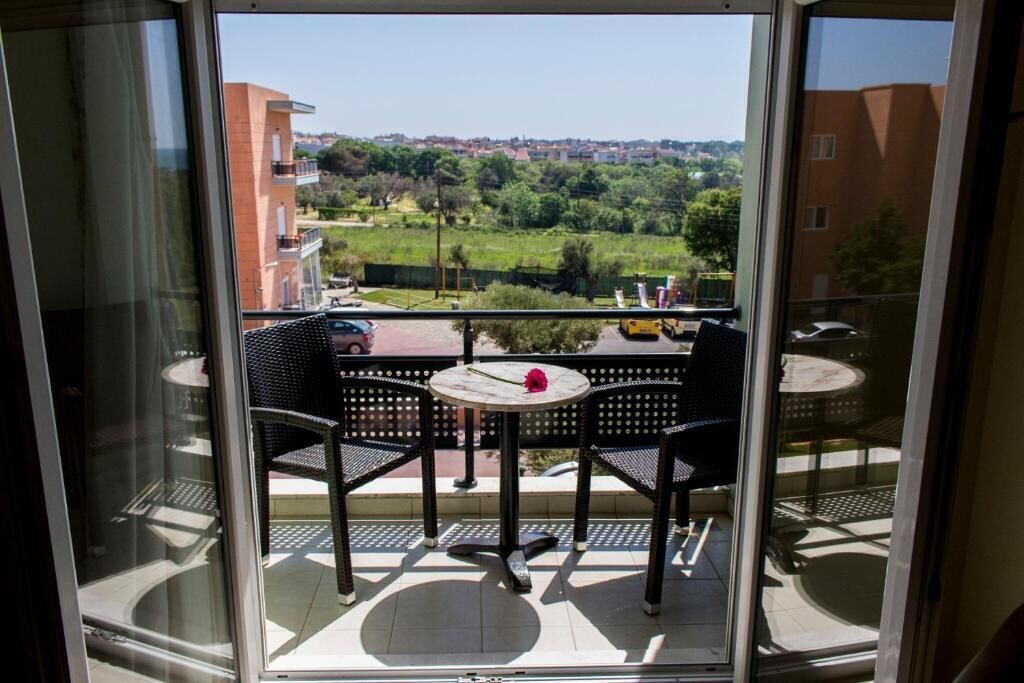A balcony with a table and chairs, offering a view of a green landscape and buildings in the distance.