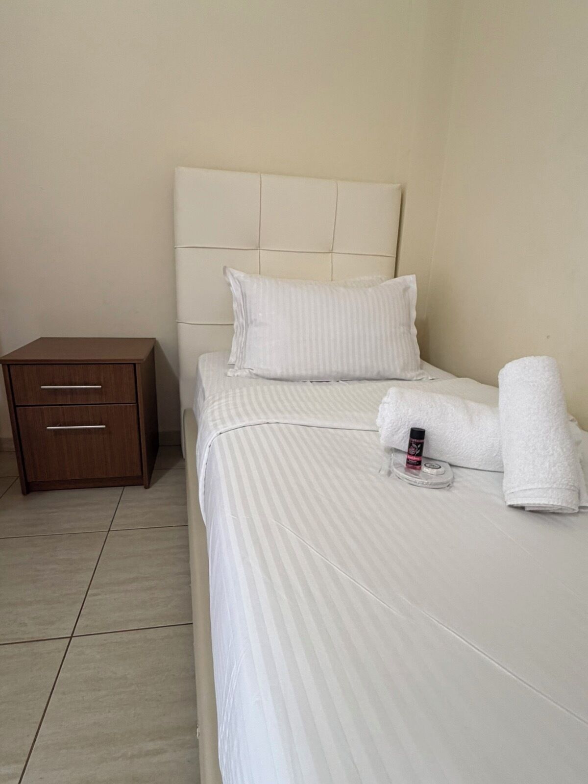 Een net opgemaakt bed met witte lakens, een houten nachtkastje met lades en een flesje lotion op het bed.
