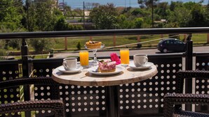 Una mesa con una porción de pastel, dos tazas y dos vasos de zumo de naranja, frente a una vista panorámica del mar y la vegetación.