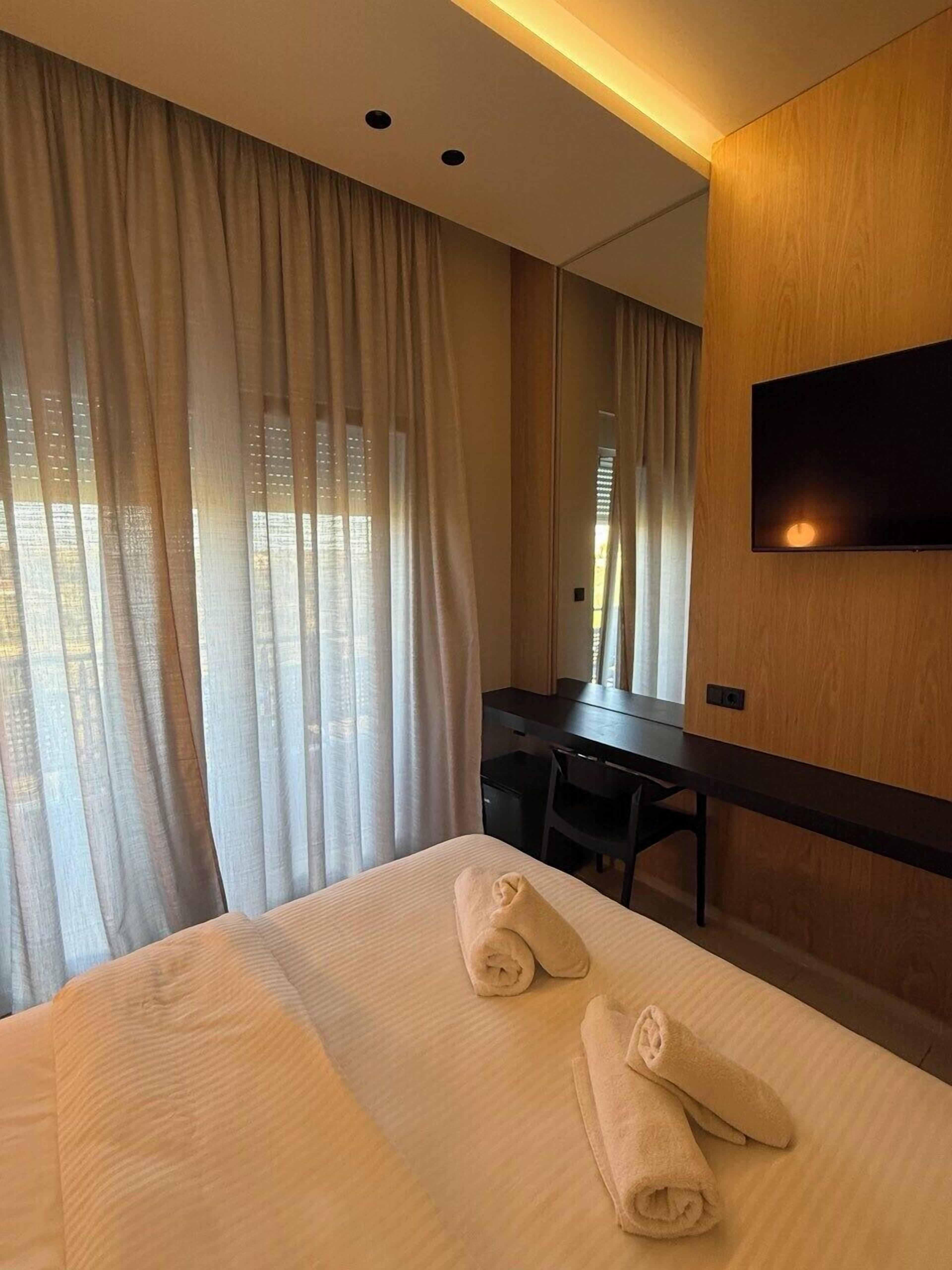 A modern hotel room with a large bed, two rolled towels, a desk, and a TV mounted on the wall.