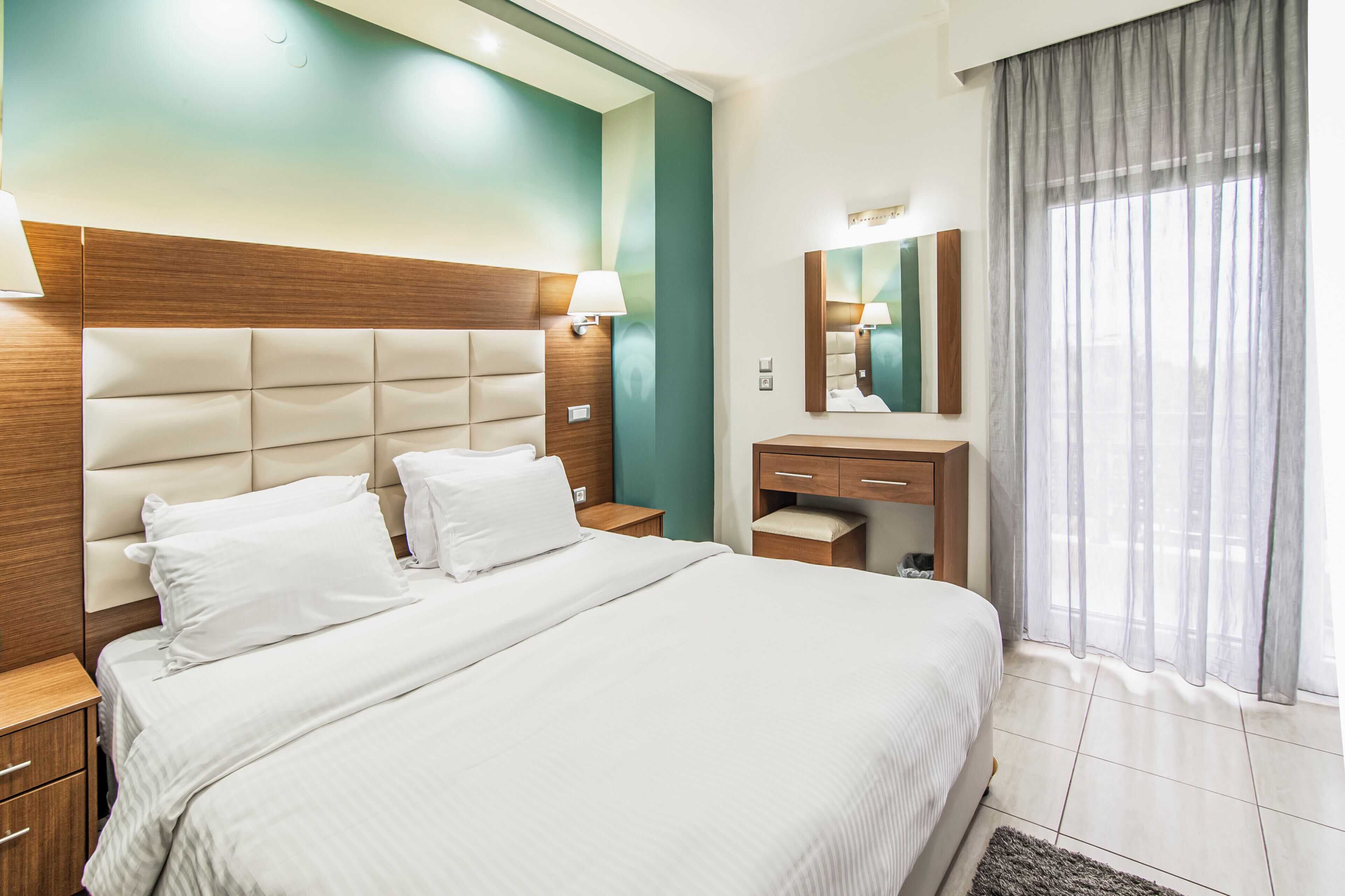 A modern hotel room with a large bed, a wooden headboard, a bedside table with a lamp, a small vanity with a mirror, and a window with sheer curtains.