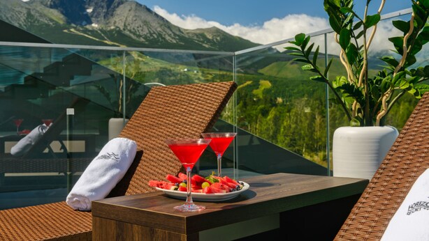 A balcony with a wooden table, two martini glasses with red drinks, a plate of watermelon and grapes, and a view of mountains. - Hotel Horizont Resort (Stara Lesna)