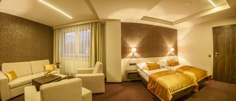 A hotel room with a sofa, armchairs, a bed, a nightstand, and a wall-mounted lamp.