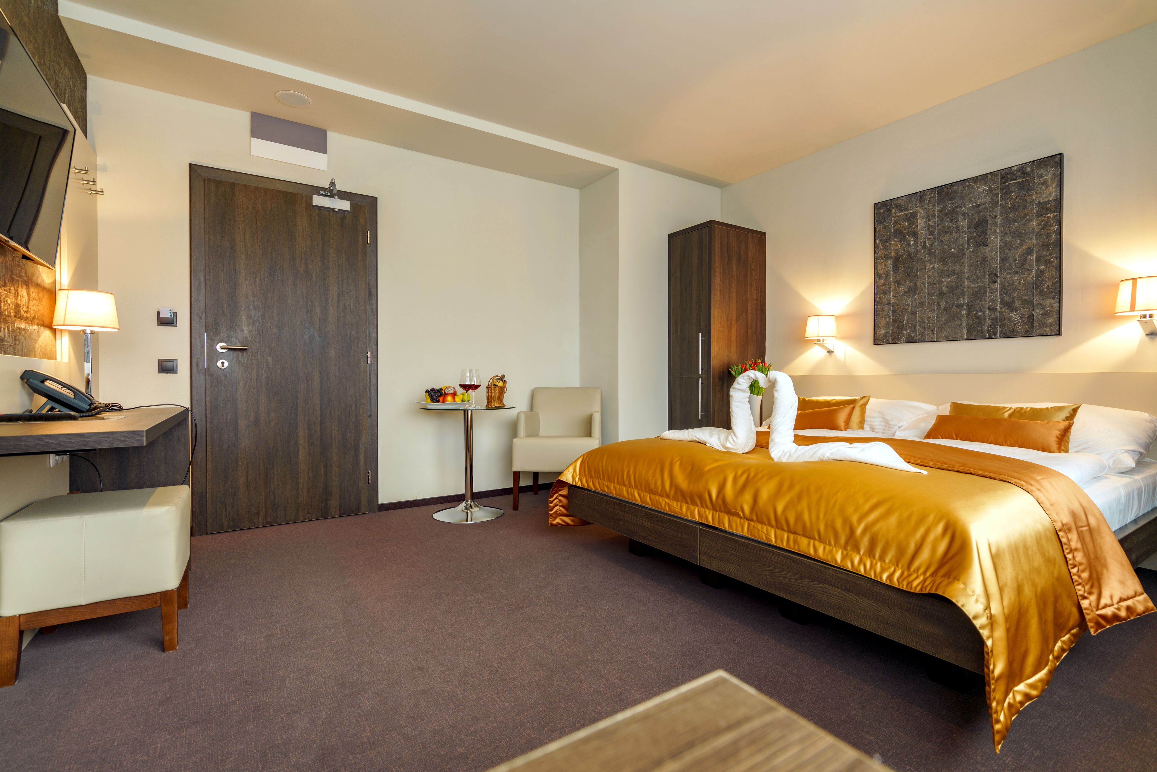 A hotel room with a bed, desk, chair, and a small table with a lamp.