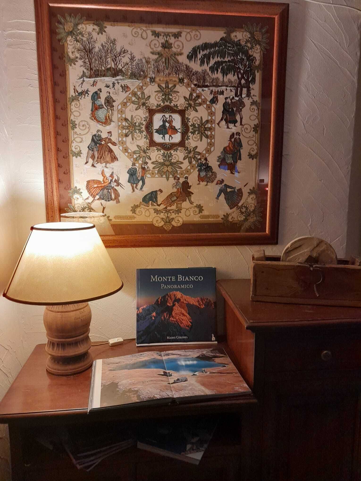 A framed historical painting with a central portrait, a book titled 'Monte Bianco Panoramico' by Mario Colonelli, and a lamp on a wooden table.
