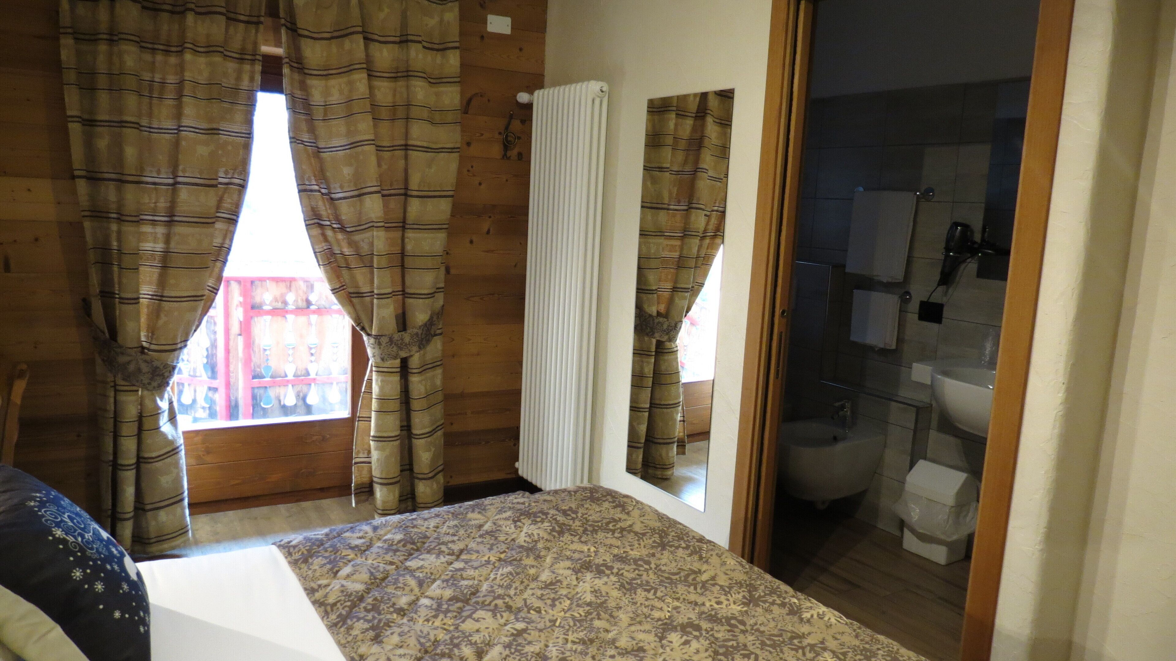 A bedroom with a bed, a window with curtains, a radiator, and a bathroom visible through an open door.