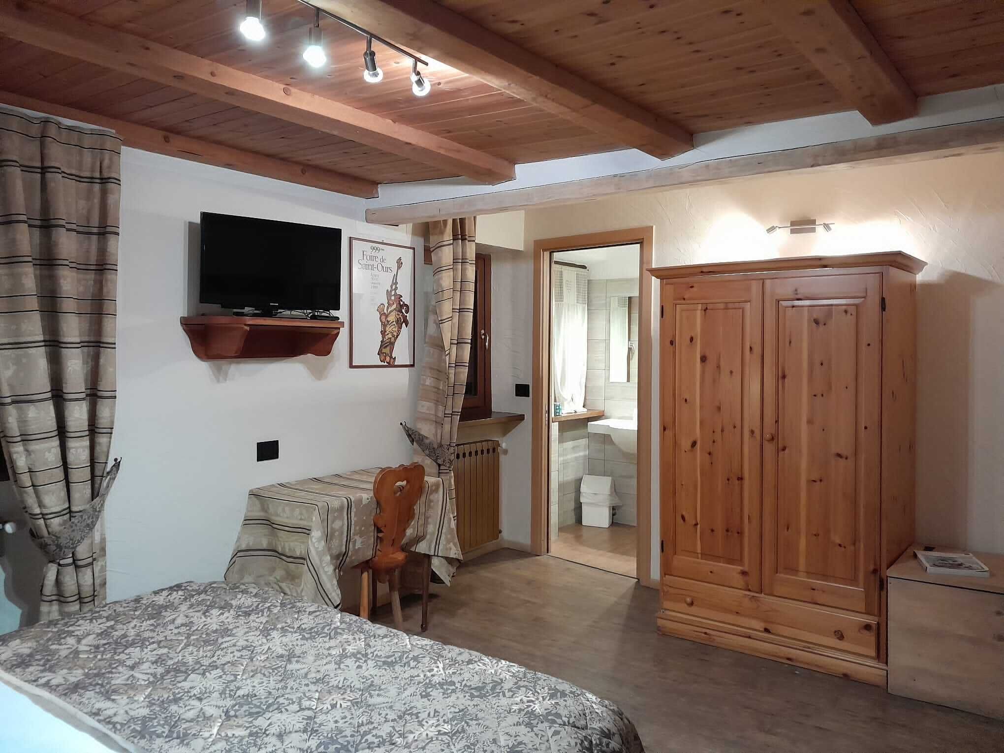 A bedroom with a bed, a wooden wardrobe, a television mounted on the wall, and a bathroom visible through an open door.