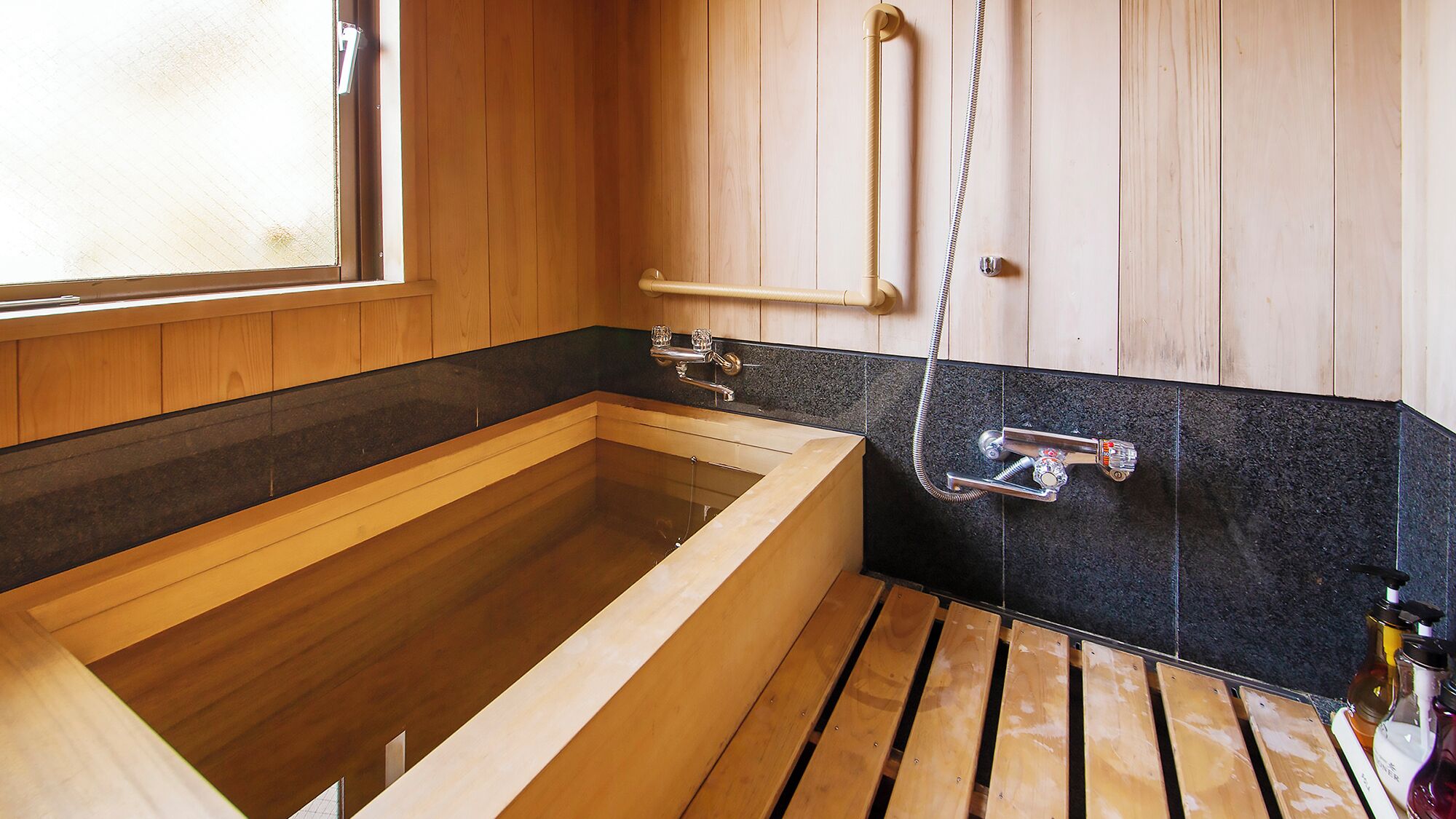 A wooden bathtub with a black tiled wall, a wooden door, and a window.
