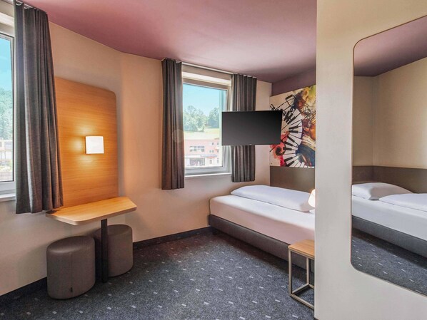 Una habitación de hotel con cama, escritorio, silla, televisión y ventana con cortinas.
