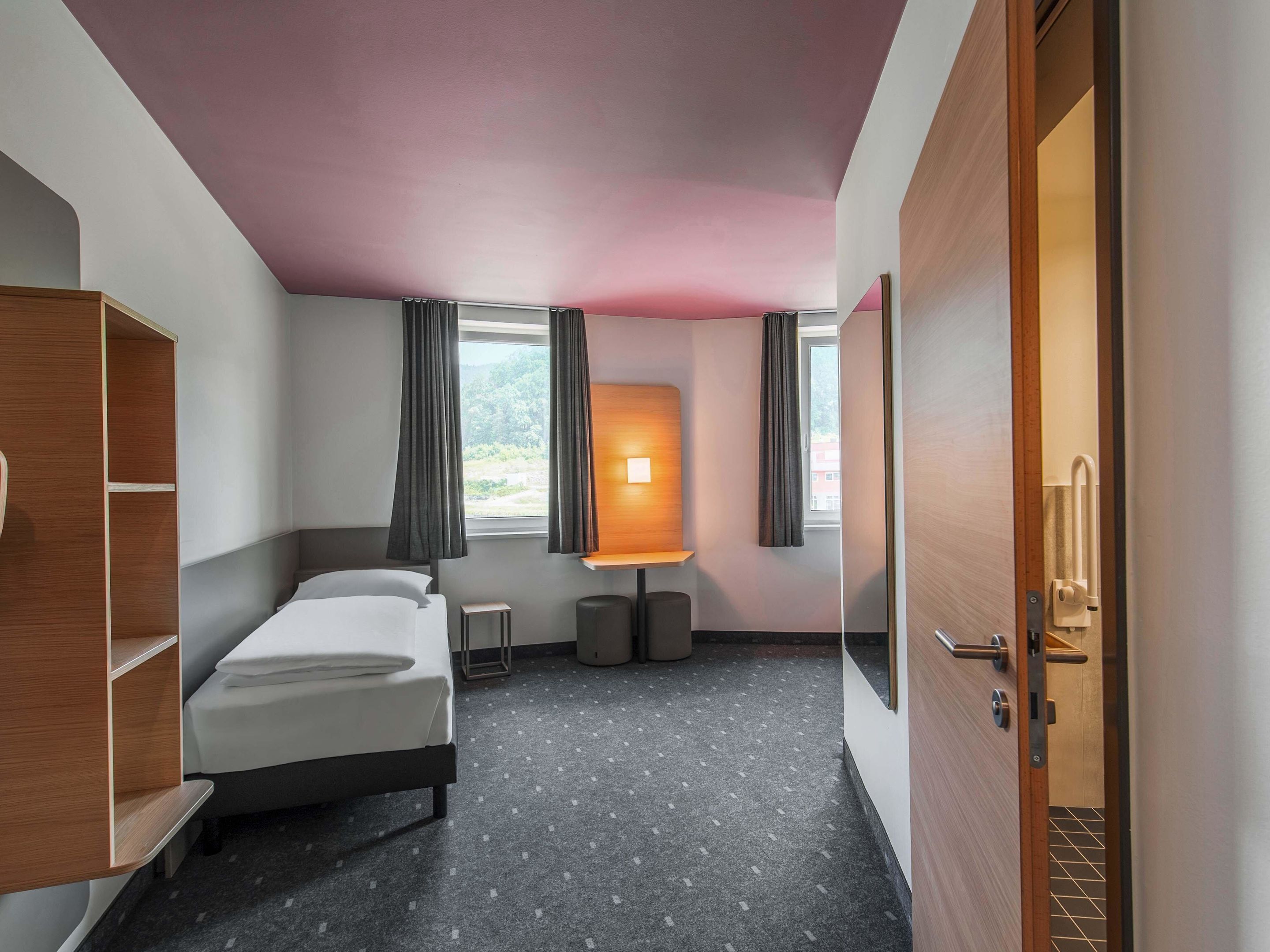 A hotel room with a bed, a desk, a chair, a lamp, and a window with curtains.