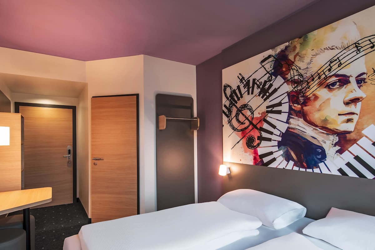 A hotel room with a bed, a desk, a chair, a door, and a wall with a large painting.