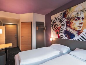 A hotel room with a bed, a desk, a chair, a door, and a wall with a large painting.